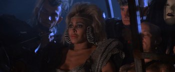 Movie still from “Mad Max Beyond Thunderdome” (1985), directed by George Miller – A woman in a silver dress with a bunch of hair on her head; Close Up shot, Over the shoulder angle