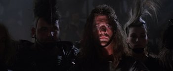 Movie still from “Mad Max Beyond Thunderdome” (1985), directed by George Miller – A man with long black hair and blue eyes is standing next to another man with long black hair and blue eyes; Close Up shot, Low angle