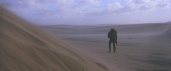 Movie still from “Mad Max Beyond Thunderdome” (1985), directed by George Miller – A man in a black jacket is standing in the sand; Extreme Wide shot, Low angle