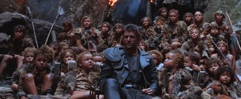 Movie still from “Mad Max Beyond Thunderdome” (1985), directed by George Miller – A man sitting in front of a group of children; Wide shot, Low angle