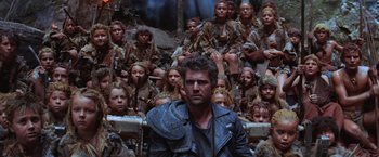 Movie still from “Mad Max Beyond Thunderdome” (1985), directed by George Miller – A group of people sitting around a man in a leather jacket; Wide shot, Low angle