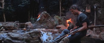 Movie still from “Mad Max Beyond Thunderdome” (1985), directed by George Miller – A man sitting on the ground next to a fire; Wide shot, Over the shoulder angle
