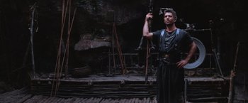 Movie still from “Mad Max Beyond Thunderdome” (1985), directed by George Miller – A man holding an axe in his hand while standing in front of a cave; Wide shot, Low angle