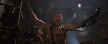 Movie still from “Mad Max Beyond Thunderdome” (1985), directed by George Miller – A man with a chain around his neck and chest is screaming; Medium shot, Low angle