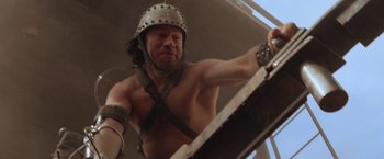 Movie still from “Mad Max Beyond Thunderdome” (1985), directed by George Miller – A shirtless man in a roman helmet is climbing a ladder; Medium shot, Low angle