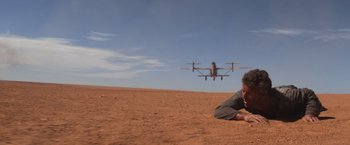 Movie still from “Mad Max Beyond Thunderdome” (1985), directed by George Miller – A man laying on the ground in front of an airplane; Wide shot, Low angle