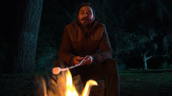 Movie still from “Mae Martin: SAP” (2023), directed by Abbi Jacobson – A man sitting in front of a fire holding a stick; Medium shot, High angle