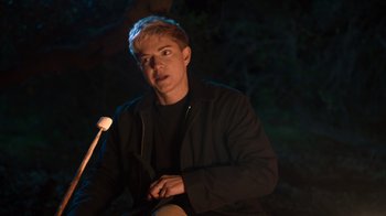 Movie still from “Mae Martin: SAP” (2023), directed by Abbi Jacobson – A man sitting in the dark holding a stick; Medium shot, Over the shoulder angle