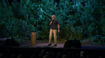 Movie still from “Mae Martin: SAP” (2023), directed by Abbi Jacobson – A man standing on a stage holding a microphone; Wide shot, High angle
