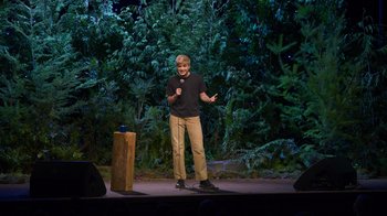 Movie still from “Mae Martin: SAP” (2023), directed by Abbi Jacobson – A man standing on a stage in front of a forest; Wide shot, High angle