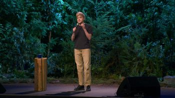 Movie still from “Mae Martin: SAP” (2023), directed by Abbi Jacobson – A man standing on a stage holding a microphone; Wide shot, High angle