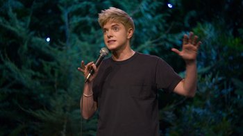 Movie still from “Mae Martin: SAP” (2023), directed by Abbi Jacobson – A man holding a microphone while standing in front of trees; Medium shot, High angle