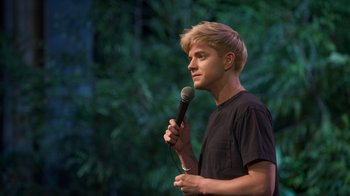 Movie still from “Mae Martin: SAP” (2023), directed by Abbi Jacobson – A man holding a microphone while standing in front of trees; Close Up shot, Over the shoulder angle