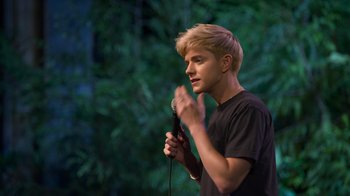 Movie still from “Mae Martin: SAP” (2023), directed by Abbi Jacobson – A man holding a microphone while standing in front of trees; Medium shot, Over the shoulder angle