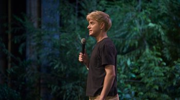 Movie still from “Mae Martin: SAP” (2023), directed by Abbi Jacobson – A man standing on a stage holding a microphone; Medium shot, High angle