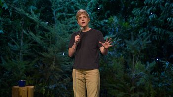 Movie still from “Mae Martin: SAP” (2023), directed by Abbi Jacobson – A man standing on a stage holding a microphone; Medium shot, High angle