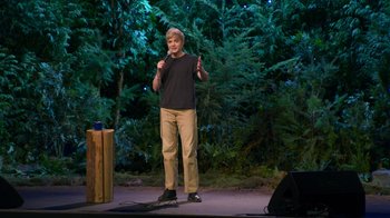Movie still from “Mae Martin: SAP” (2023), directed by Abbi Jacobson – A man standing on a stage in front of trees; Medium shot, High angle