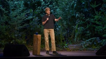 Movie still from “Mae Martin: SAP” (2023), directed by Abbi Jacobson – A man standing on a stage holding a microphone; Medium shot, High angle