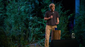 Movie still from “Mae Martin: SAP” (2023), directed by Abbi Jacobson – A man standing on a stage holding a microphone; Medium shot, Over the shoulder angle