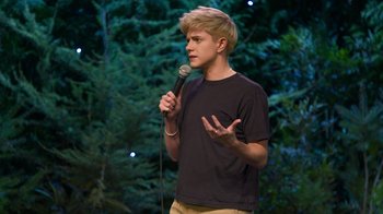 Movie still from “Mae Martin: SAP” (2023), directed by Abbi Jacobson – A man holding a microphone while standing in front of a forest; Medium shot, High angle