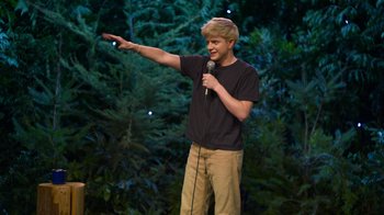 Movie still from “Mae Martin: SAP” (2023), directed by Abbi Jacobson – A man standing on a stage holding a microphone; Medium shot, High angle