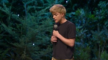 Movie still from “Mae Martin: SAP” (2023), directed by Abbi Jacobson – A man holding a microphone in front of trees; Medium shot, Over the shoulder angle