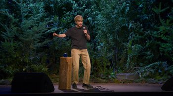 Movie still from “Mae Martin: SAP” (2023), directed by Abbi Jacobson – A man standing on a stage holding a microphone; Wide shot, High angle