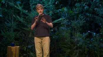 Movie still from “Mae Martin: SAP” (2023), directed by Abbi Jacobson – A man standing on a stage holding a microphone; Wide shot, High angle