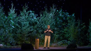 Movie still from “Mae Martin: SAP” (2023), directed by Abbi Jacobson – A man standing on a stage in front of a bunch of trees; Wide shot, Over the shoulder angle