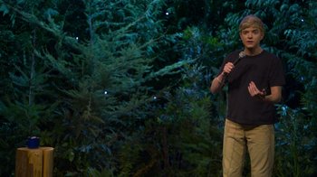 Movie still from “Mae Martin: SAP” (2023), directed by Abbi Jacobson – A man standing in front of a tree with a microphone; Medium shot, High angle