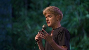 Movie still from “Mae Martin: SAP” (2023), directed by Abbi Jacobson – A man holding a microphone in front of trees; Close Up shot, High angle
