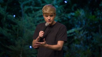 Movie still from “Mae Martin: SAP” (2023), directed by Abbi Jacobson – A man holding a microphone while standing in front of trees; Close Up shot, Over the shoulder angle
