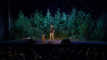 Movie still from “Mae Martin: SAP” (2023), directed by Abbi Jacobson – A man standing on a stage in front of a crowd; Extreme Wide shot, High angle