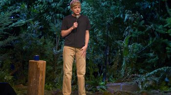 Movie still from “Mae Martin: SAP” (2023), directed by Abbi Jacobson – A man standing on a stage holding a microphone; Wide shot, High angle