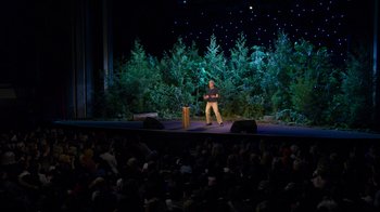 Movie still from “Mae Martin: SAP” (2023), directed by Abbi Jacobson – A man standing on a stage in front of an audience; Extreme Wide shot, High angle