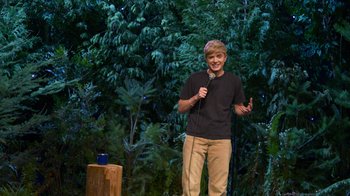 Movie still from “Mae Martin: SAP” (2023), directed by Abbi Jacobson – A young man standing on a stage holding a microphone; Wide shot, High angle
