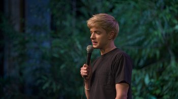 Movie still from “Mae Martin: SAP” (2023), directed by Abbi Jacobson – A man holding a microphone while standing on a stage; Medium shot, High angle