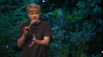 Movie still from “Mae Martin: SAP” (2023), directed by Abbi Jacobson – A person holding a microphone on a stage; Medium shot, Over the shoulder angle