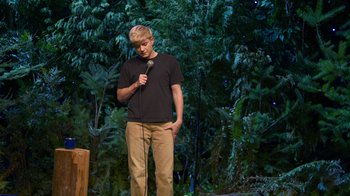 Movie still from “Mae Martin: SAP” (2023), directed by Abbi Jacobson – A man standing on a stage holding a microphone; Wide shot, Over the shoulder angle