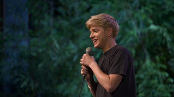 Movie still from “Mae Martin: SAP” (2023), directed by Abbi Jacobson – A young man holding a microphone while standing on a stage; Medium shot, High angle