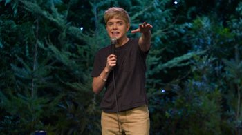 Movie still from “Mae Martin: SAP” (2023), directed by Abbi Jacobson – A young man standing on a stage holding a microphone; Medium shot, Over the shoulder angle