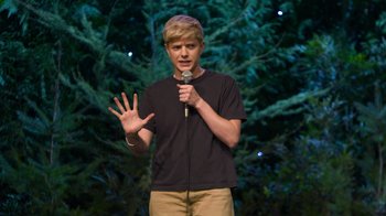 Movie still from “Mae Martin: SAP” (2023), directed by Abbi Jacobson – A man standing on a stage holding a microphone; Medium shot, High angle