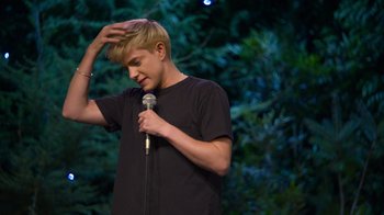 Movie still from “Mae Martin: SAP” (2023), directed by Abbi Jacobson – A man standing on a stage holding a microphone; Medium shot, Over the shoulder angle