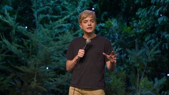 Movie still from “Mae Martin: SAP” (2023), directed by Abbi Jacobson – A young man holding a microphone while standing in front of a tree; Medium shot, High angle