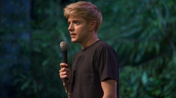 Movie still from “Mae Martin: SAP” (2023), directed by Abbi Jacobson – A man holding a microphone in front of trees; Close Up shot, Over the shoulder angle