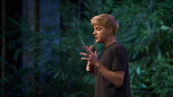 Movie still from “Mae Martin: SAP” (2023), directed by Abbi Jacobson – A man standing on a stage holding a microphone; Medium shot, Low angle