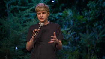 Movie still from “Mae Martin: SAP” (2023), directed by Abbi Jacobson – A man standing on a stage holding a microphone; Medium shot, High angle