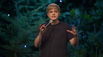 Movie still from “Mae Martin: SAP” (2023), directed by Abbi Jacobson – A young man holding a microphone while standing in front of trees; Medium shot, Low angle