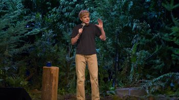 Movie still from “Mae Martin: SAP” (2023), directed by Abbi Jacobson – A man standing on a stage holding a microphone; Wide shot, High angle