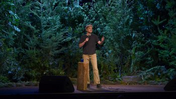 Movie still from “Mae Martin: SAP” (2023), directed by Abbi Jacobson – A man standing on a stage in front of trees; Wide shot, High angle